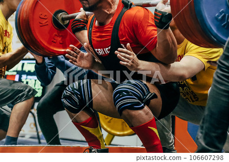 male athlete squat in powerlifting, knees are wrapped with an elastic bandage, coach hands are on safety net male athlete squat in powerlifting, knees are wrapped with an elastic bandage, coach hands are on safety net 106607298