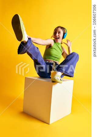 Cheerful, positive, smiling young woman in stylish clothes listening to music in headphones against yellow studio background 106607306