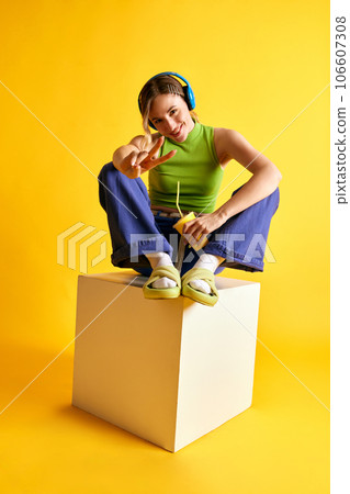 Cheerful, positive, smiling young woman in stylish clothes listening to music in headphones against yellow studio background Cheerful, positive, smiling young woman in stylish clothes listening to music in headphones against yellow studio background 106607308