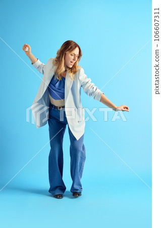 Smiling, delightful young woman listening to music in earphones, dancing and singing against blue studio background. Enjoyment Smiling, delightful young woman listening to music in earphones, dancing and singing against blue studio background. Enjoyment 106607311