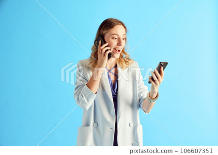 Young woman, employee talking on mobile phone and looking at another phone against blue studio background 106607351