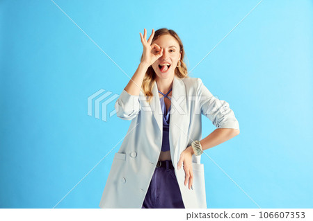 Okey. Cheerful, positive young women in blazer holding okay hand gesture over eye against blue studio background 106607353