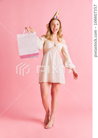 Full-length image of cheerful, happy young woman in tender dress walking with shopping bags, presents against pink studio background 106607357