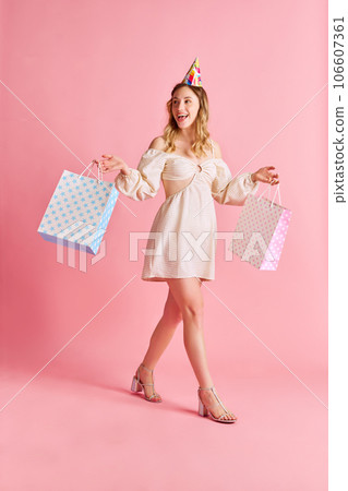 Full-length of smiling, happy young girl in cute dress walking with colorful bags against pink studio background. Presents Full-length of smiling, happy young girl in cute dress walking with colorful bags against pink studio background. Presents 106607361