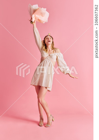 Full-length image of happy emotional young woman in cute dress standing with flowers and laughing against pink studio background Full-length image of happy emotional young woman in cute dress standing with flowers and laughing against pink studio background 106607362