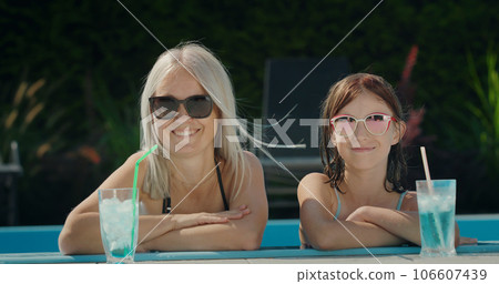 Portrait of a mother with her daughter, relaxing in the pool. Cooling drinks are next to them. Looking at the camera 106607439