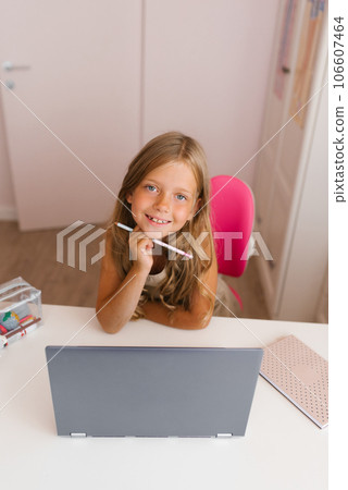 Little girl using a laptop at home for online education and home schooling, looks at the camera with a smile at home and holds a pen in her hand. Distance learning for children 106607464