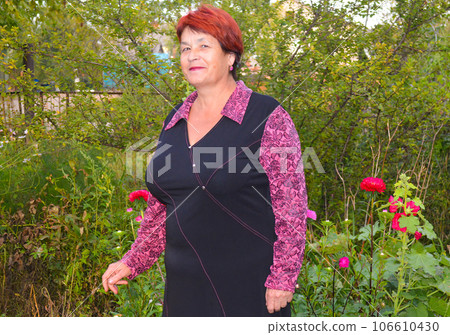 Elderly good-looking woman in dress smiling and standing near beautiful pink and red asters. 106610430