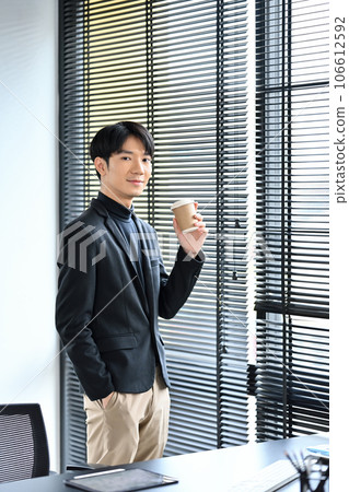 Handsome young businessman holding disposable cup standing in office and looking at camera Handsome young businessman holding disposable cup standing in office and looking at camera 106612592