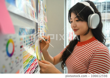Pretty young creative woman listening to music in headphone and working with color palette at graphic studio Pretty young creative woman listening to music in headphone and working with color palette at graphic studio 106612598