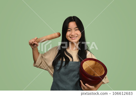 Lovely young asian girl wearing apron holding mortar and pestle isolated on green background 106612683