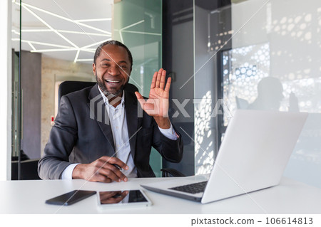 Portrait of successful mature african american boss, man smiling and looking at camera sitting at desk inside office, businessman waving hand gesture of greeting and friendship. 106614813