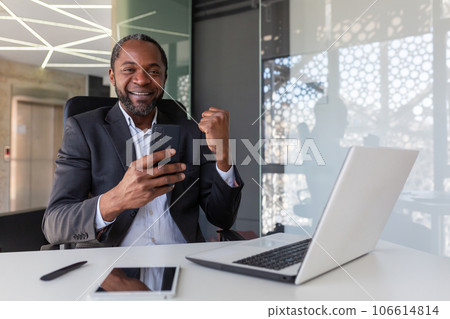 Portrait of successful winner at workplace inside office, African American man smiling and looking at camera, businessman holding phone, received online notification of winning message. 106614814