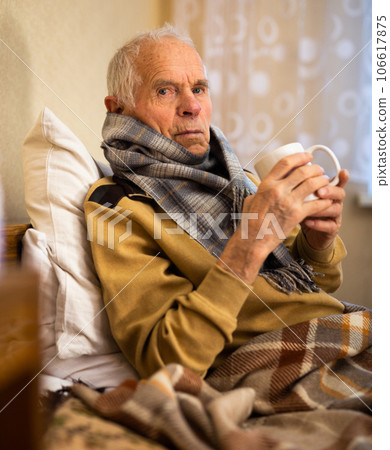 Old man wrapped in blanket has cold and drinks hot infusion 106617875