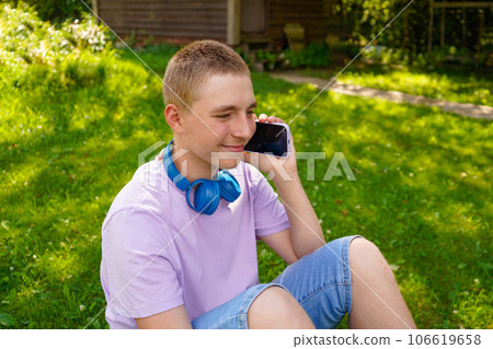 Young teen speaks on mobile phone sitting on the green grass. wearing headphones 106619658