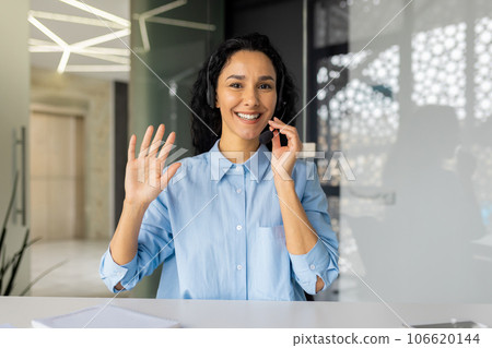 Webcam view, woman with headset phone smiling and looking at camera, waving hand gesture of friendship and greeting, online customer support worker at workplace inside office smiling. 106620144