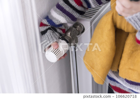 A small child warmly dressed in a sweater and a hat turns the tap on the radiator.  106620718