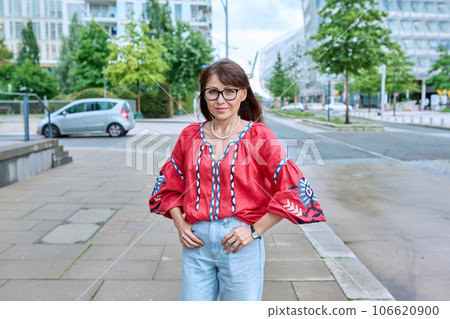 Mature smiling woman in embroidered shirt looking at camera, on street of modern city Mature smiling woman in embroidered shirt looking at camera, on street of modern city 106620900