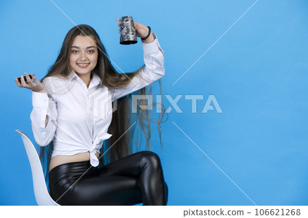 Cheerful girl in white blouse, flipping paper cup upside down, while sitting on chair. Smiling caucasian woman holding empty cup, while looking at camera, isolated on blue background. Concept of fun. 106621268