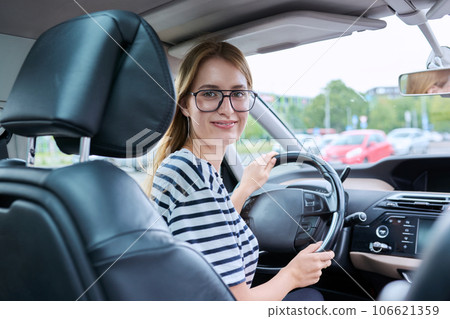 Teenage girl driver in glasses sitting behind wheel of car, looking at camera 106621359