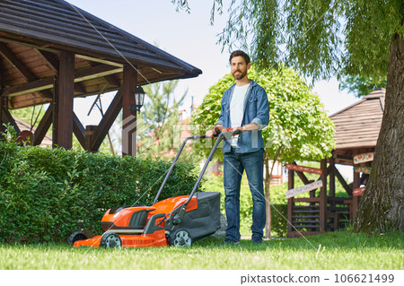Smiling male landscaper in denim shirt trimming overgrown lawn with lawn mover at summer day. Low angle view of happy bearded guy using electric mower, while smiling at camera. Concept of gardening. Smiling male landscaper in denim shirt trimming overgrown lawn with lawn mover at summer day. Low angle view of happy bearded guy using electric mower, while smiling at camera. Concept of gardening. 106621499