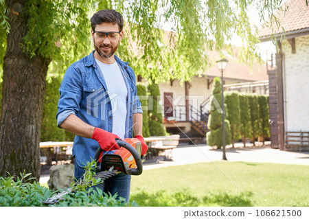 Brunette male landscaper in protective glasses cutting bush with hedge trimmer outside. Front view of professional gardener using modern equipment, while taking care of plants. Concept of workplace. Brunette male landscaper in protective glasses cutting bush with hedge trimmer outside. Front view of professional gardener using modern equipment, while taking care of plants. Concept of workplace. 106621500