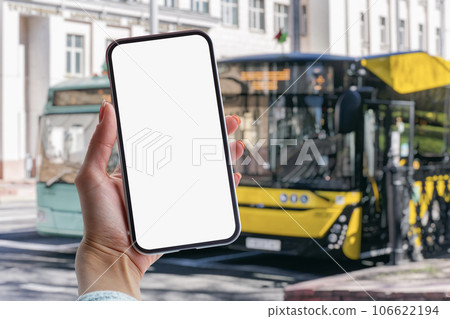 Girl holds a mock-up of a smartphone with a white screen in her hands at a bus stop against the backdrop of a bus. 106622194