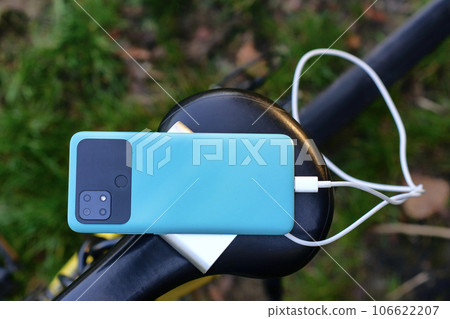 A portable charger charges a smartphone on the background of a bicycle and nature. Power Bank on a journey to charge gadgets. 106622207
