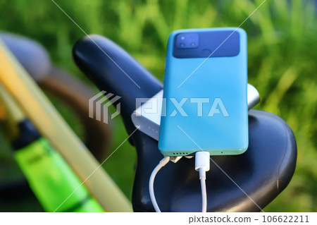 A portable charger charges a smartphone on the background of a bicycle and nature. Power Bank on a journey to charge gadgets. 106622211