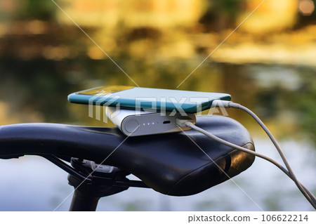 A portable charger charges a smartphone on the background of a bicycle and nature. Power Bank on a journey to charge gadgets. A portable charger charges a smartphone on the background of a bicycle and nature. Power Bank on a journey to charge gadgets. 106622214
