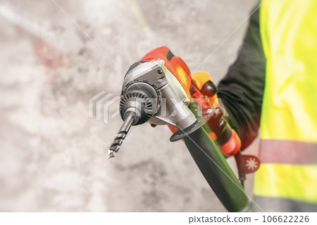 Male worker holds a close-up electronic drill in his hands against the background of a concrete wall. 106622226