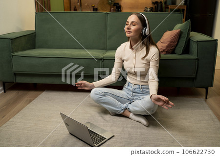 Young brunette woman wearing headphones practices yoga online while studying a laptop on a yoga mat at home. 106622730
