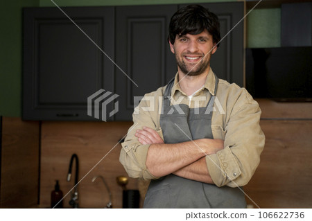 Handsome smiling young man in an apron looking at the camera. Portrait of a happy laid-back guy in an apron standing in the kitchen Handsome smiling young man in an apron looking at the camera. Portrait of a happy laid-back guy in an apron standing in the kitchen 106622736