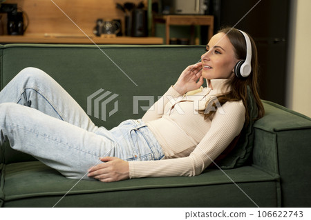 Beautiful young woman lying on a green sofa with headphones and eyes closed, relaxing listening to music 106622743
