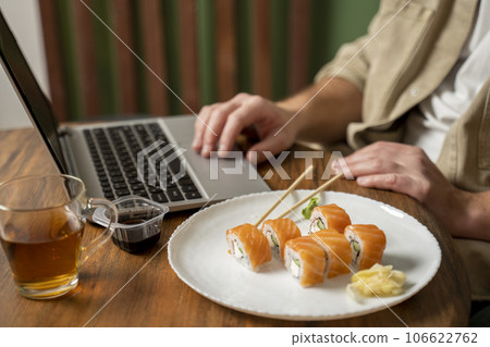 Close-up of a young handsome man wearing beige shirt working on a laptop, eating sushi with salmon 106622762