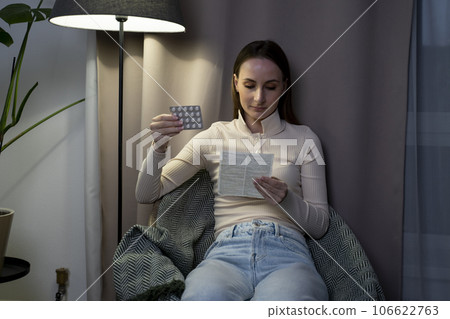 A woman taking pills and reading instructions, medical rehabilitation and a pharmacy box with pills in the living room of the house. 106622763
