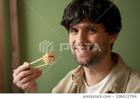 Young handsome man wearing beige shirt eating sushi roll with salmon using chopsticks 106622764
