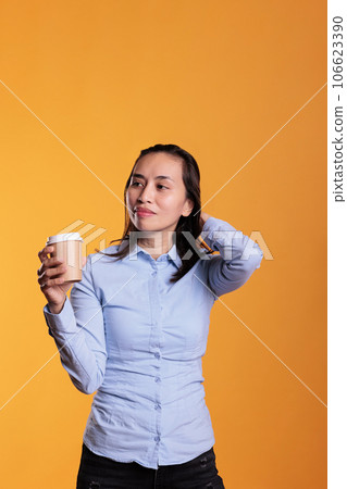 Filipino woman posing in studio with cup of coffee, enjoying drinking hot beverage over yellow background. Positive adult holding papercup using drink to have energy and refreshment. 106623390