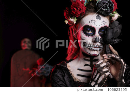Spooky santa muerte lady posing with black roses, acting like holy goddess of death on day of the dead mexican holiday. Celebrating halloween ritual with la cavalera catrina skull body art. 106623395