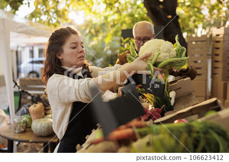 Elderly man and young vendor putting colorful products on farmers market stand, selling healthy grown fruits and veggies. Team of farmers serving customers with organic bio local produce. 106623412