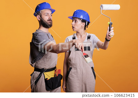 Young construction workers posing with painting tools, standing over yellow background. Team of constructors holding paintbrush and roller brush to work on refurbishment project in studio. 106623415