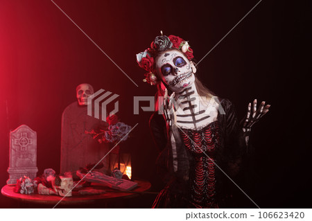 Scary woman talking on mobile phone call, wearing halloween make up and flowers crown in studio. La cavalera catrina using smartphone line on dios de los muertos mexican tradition. Scary woman talking on mobile phone call, wearing halloween make up and flowers crown in studio. La cavalera catrina using smartphone line on dios de los muertos mexican tradition. 106623420