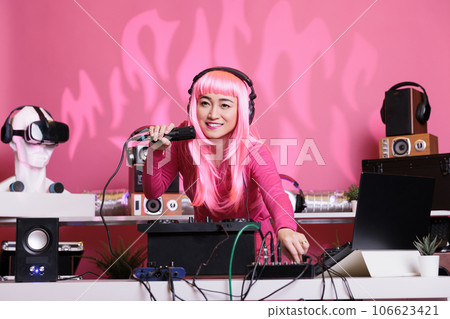 Artist with pink hair mixing techno song with electronic using professional turntables while enjoying talking with fans, having fun together during night time in club. Woman enjoying performing music 106623421