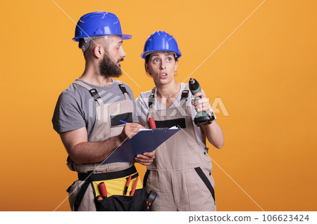 Team of construction workers taking notes on clipboard and checking renovation measurement, holding drill gun. Man and woman renovators looking at papers or files, standing over yellow background. Team of construction workers taking notes on clipboard and checking renovation measurement, holding drill gun. Man and woman renovators looking at papers or files, standing over yellow background. 106623424