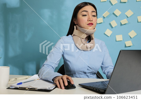 Corporate employee wearing medical collar for injury while working on reports, preparing paperwork for next meeting. Injured female entrepreneur concentrating on finishing project at work station. 106623451