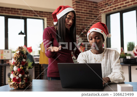 Asian and african american women working together on laptop in christmas decorated office. Project manager in santa hat helping colleague while using computer at festive workplace Asian and african american women working together on laptop in christmas decorated office. Project manager in santa hat helping colleague while using computer at festive workplace 106623499