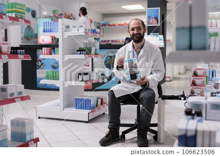 Smiling pharmacist standing on chair in pharmacy holding medical flyer to promote health care support service. Drugstore filled with pharmaceutical products, vitamins and drugs Smiling pharmacist standing on chair in pharmacy holding medical flyer to promote health care support service. Drugstore filled with pharmaceutical products, vitamins and drugs 106623506