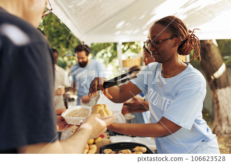 Detailed image of black woman at a food drive sharing free warm meals to poor caucasian homeless person. At local center group of volunteers feed and support the hungry and underprivileged. 106623528