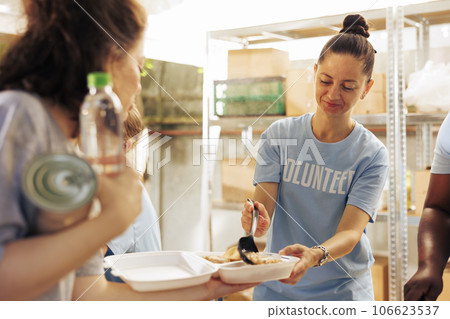 Young woman providing assistance, serving warm meals and supporting homeless people and refugees in need. Image showing female charity worker handing out free food to the less privileged. Young woman providing assistance, serving warm meals and supporting homeless people and refugees in need. Image showing female charity worker handing out free food to the less privileged. 106623537