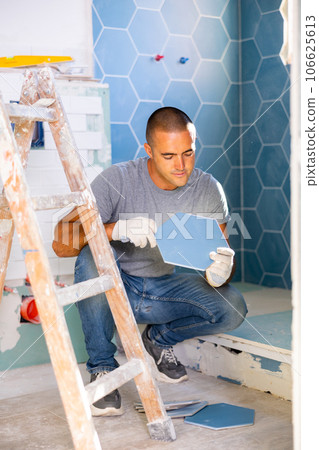 Portrait of concentrated young man sitting squat and making renovation repair. Tiler installing tile on wall 106625613